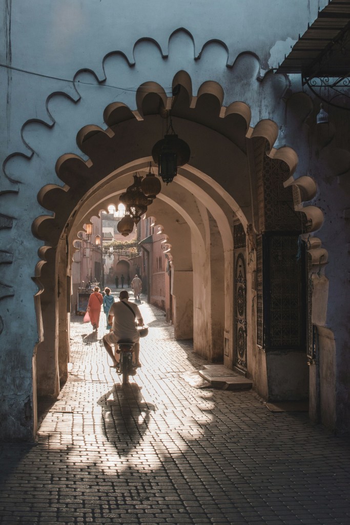 A person riding a bicycle through an archway in a narrow street with cobblestones, illuminated by soft sunlight. In the background, various people are walking, surrounded by traditional architecture.