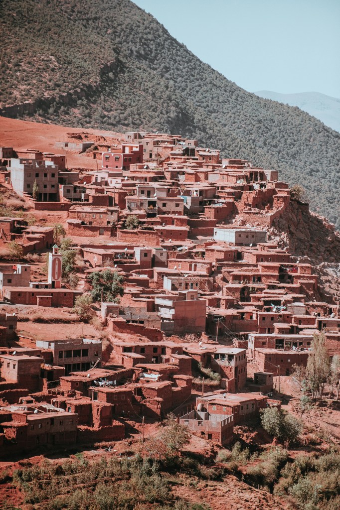 Aerial view of a hillside village with buildings made of red earth, surrounded by lush greenery and mountains in the background.