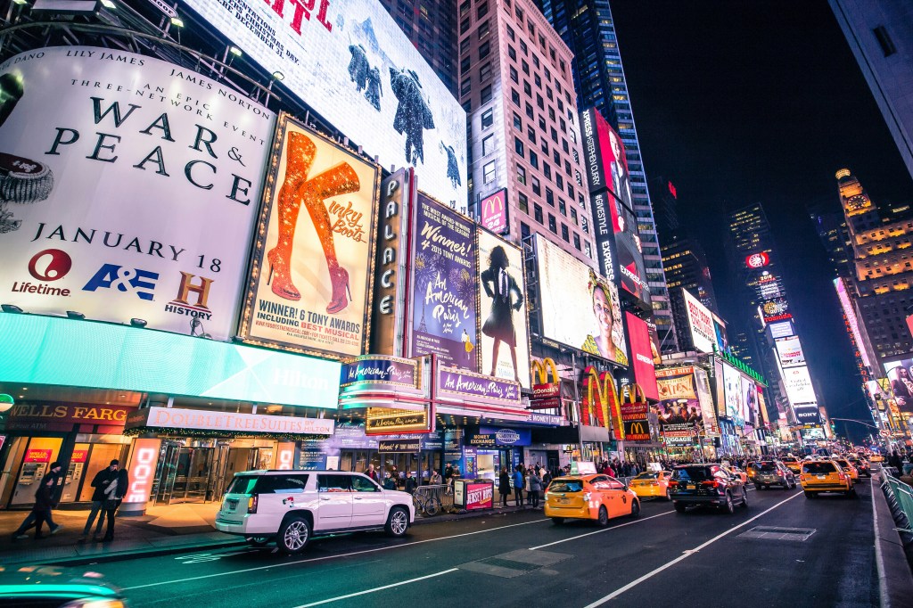 A bustling night scene in Times Square, showcasing numerous bright advertisements and billboards, including 'War & Peace' and 'Kinky Boots,' with cars and taxis driving on the street.