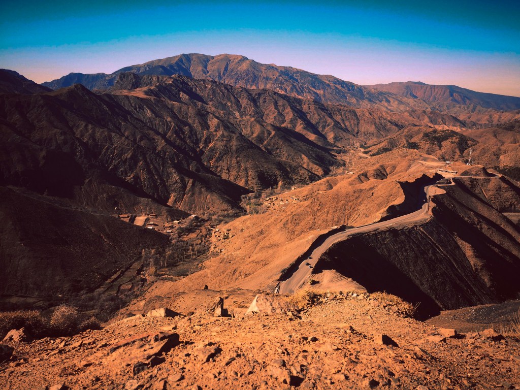 A panoramic view of rugged, mountainous terrain featuring steep slopes and a winding road through arid landscape under a clear blue sky.