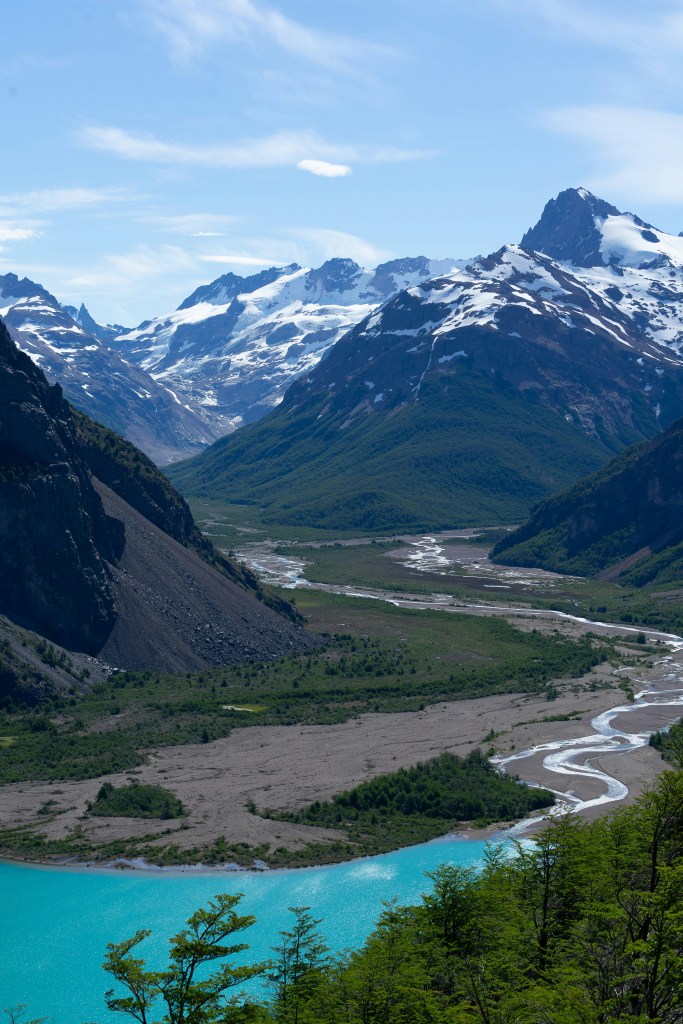 Scenic view of mountains with snow caps surrounding a turquoise river and green valley, under a clear blue sky.