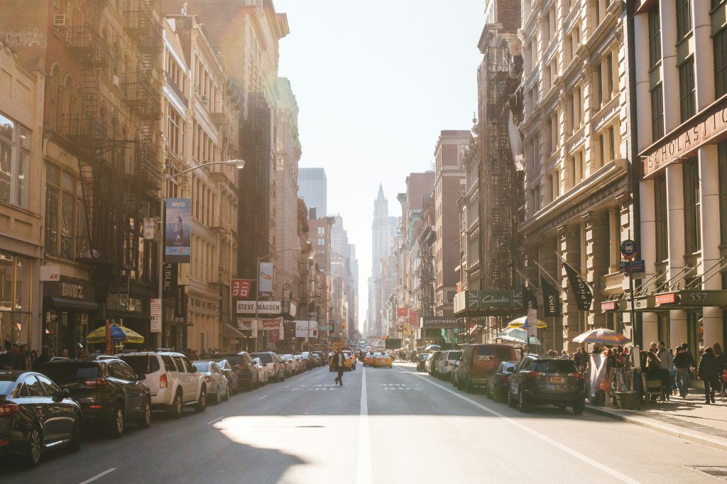 A busy city street lined with cars and shops, featuring tall buildings and sunlight streaming down the road.