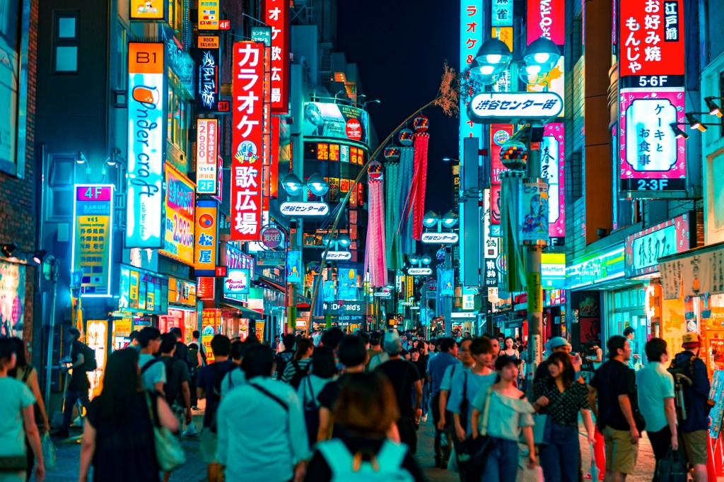 A busy street scene at night in a city, filled with colorful neon signs and large advertisements. Many people are walking along the sidewalk, creating a vibrant and lively atmosphere.