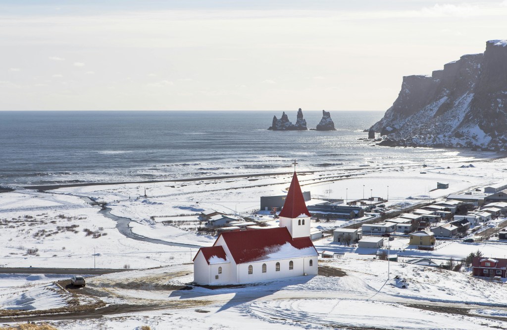 A small white church with a red roof stands in a snowy landscape overlooking the ocean, with cliffs and rock formations in the background.