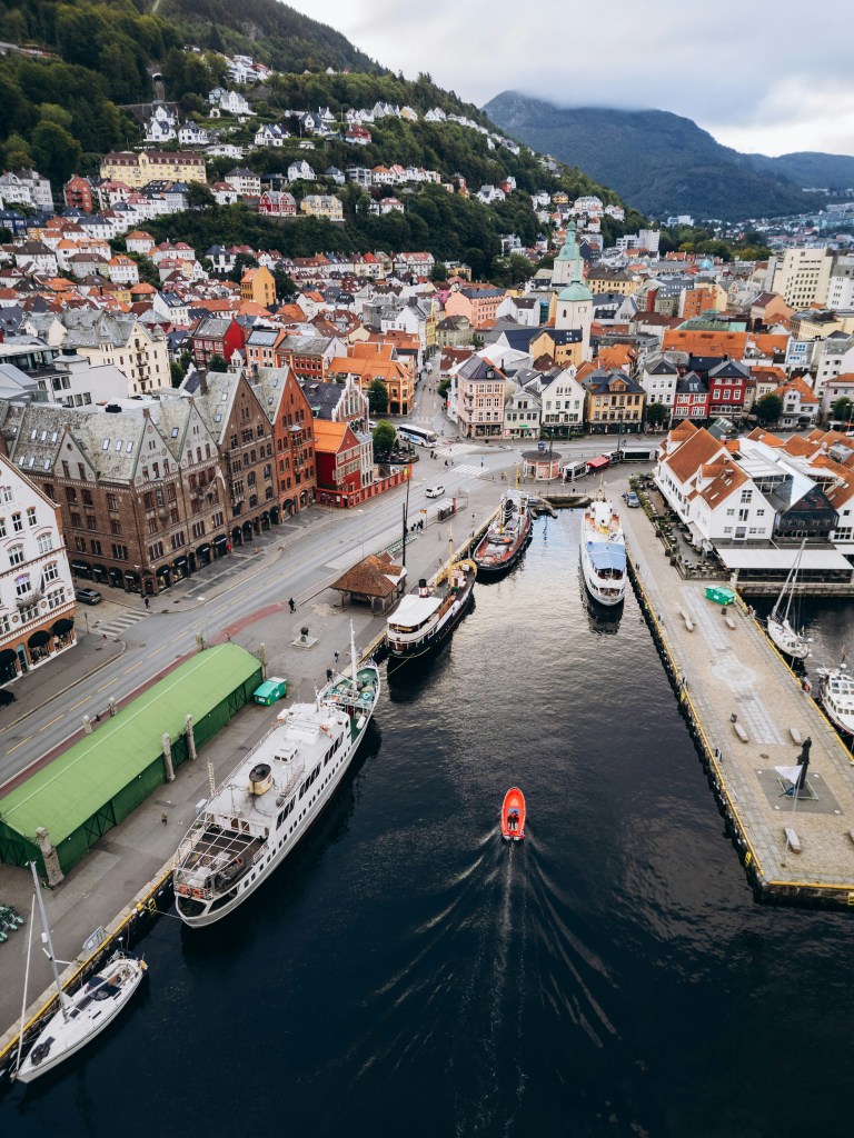 Aerial view of a colorful waterfront city, showcasing a mix of traditional and modern architecture, boats docked along the harbor, and lush green hills in the background.