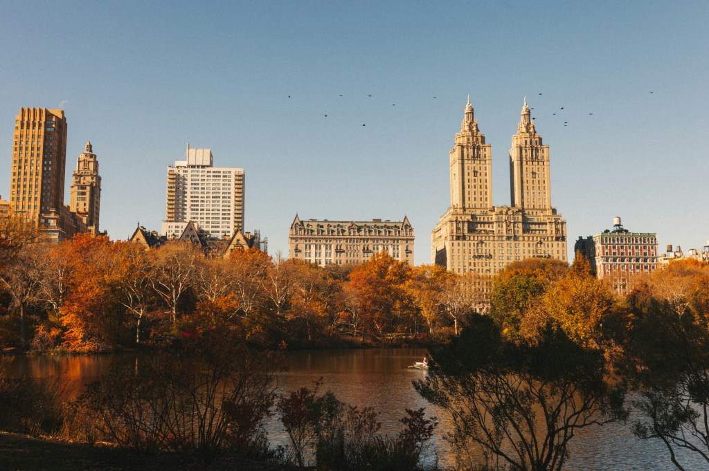 Autumn view of Central Park with colorful trees and skyline featuring tall buildings against a clear blue sky.