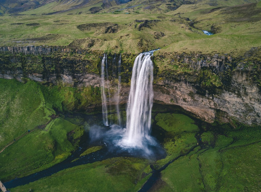 Aerial view of Seljalandsfoss waterfall cascading over a rocky cliff surrounded by lush green hills and vegetation in Iceland.