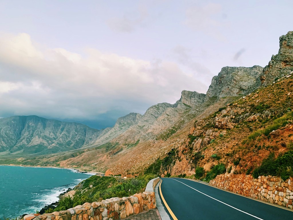 A winding road alongside a rocky coastline with mountains in the background and a cloudy sky.