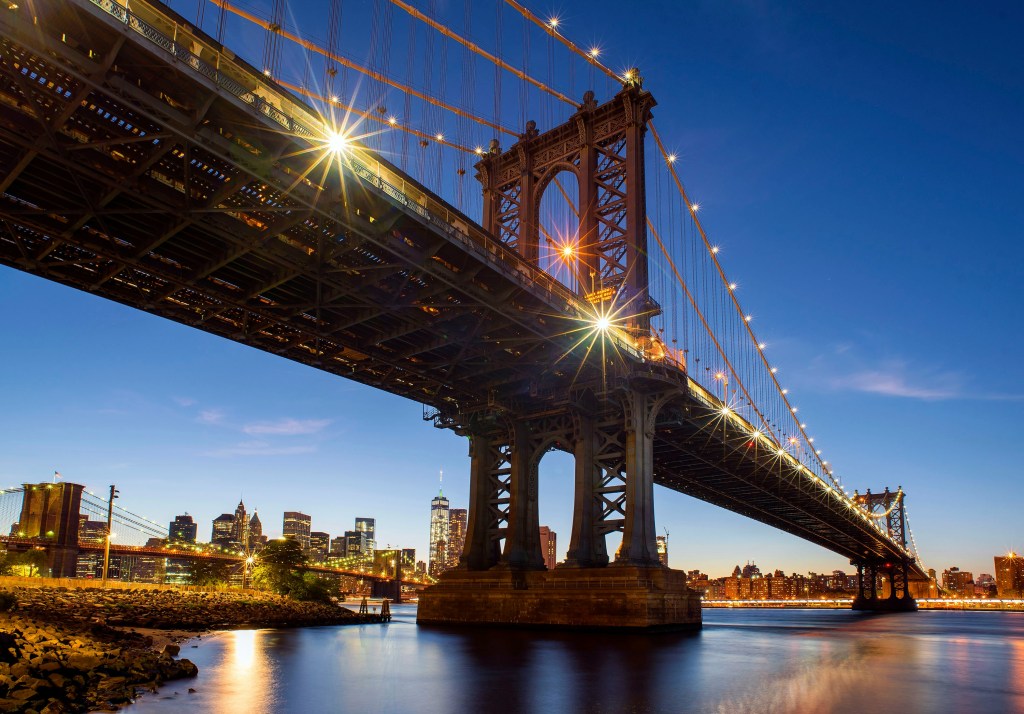 A nighttime view of the Manhattan Bridge, featuring sparkling lights and reflections on the water, with the skyline of New York City in the background.