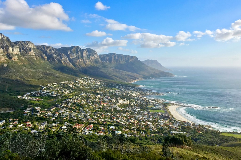 A panoramic view of a coastal town nestled between lush green hills and rugged mountains, with the ocean visible in the background under a partly cloudy sky.